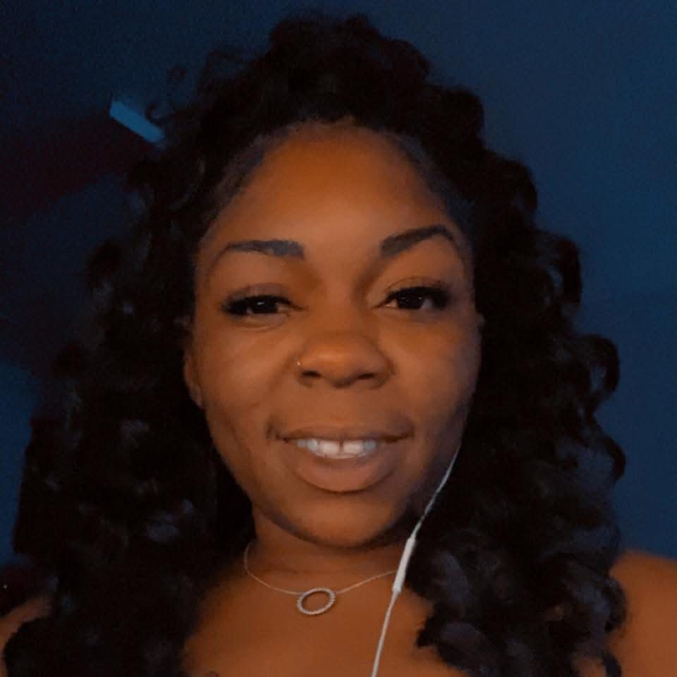 Person with long curly hair wearing a necklace with a circular pendant, photographed against a dark background.